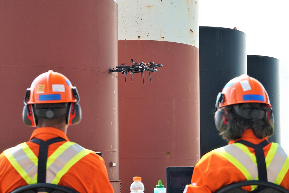 Drone controlled by two workers in safety gear, used to perform a wall thickness inspection of an oil refinery storage tank